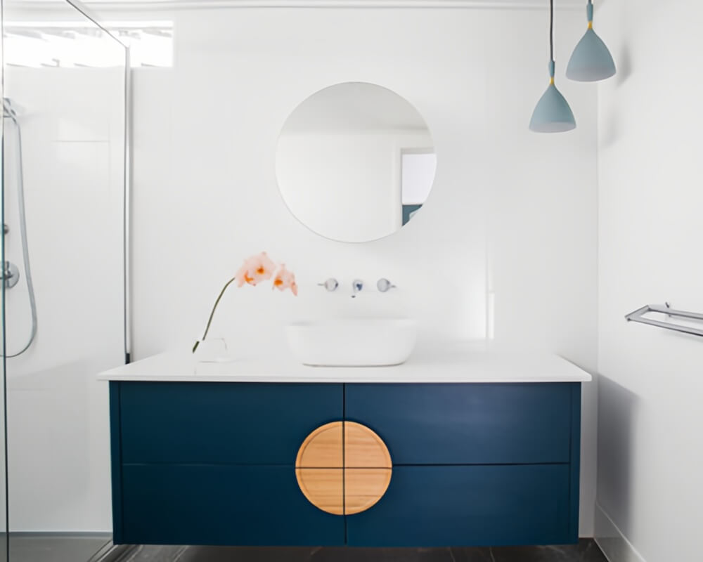 Navy blue bathroom vanity with round white vessel sink, frameless round mirror, wooden knobs, glass shower partition, and soft blue pendant lights