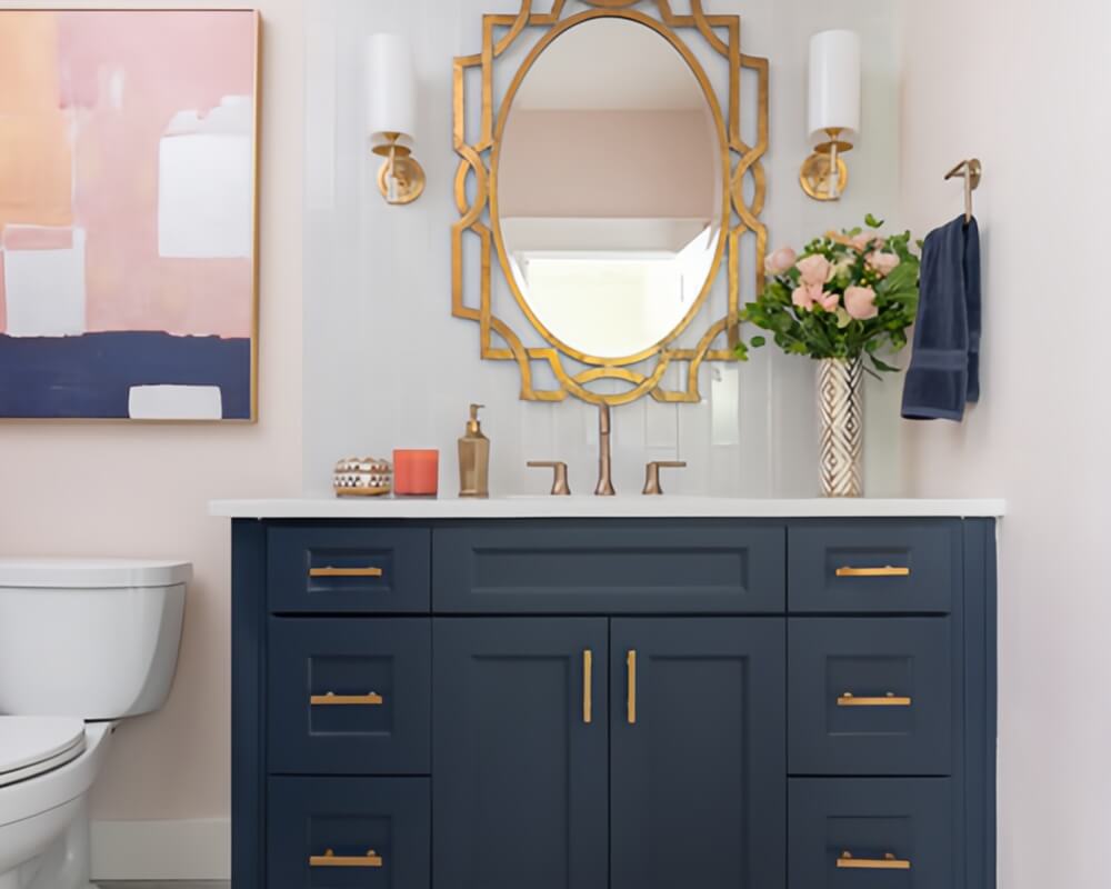 Navy blue bathroom vanity with gold handles, white marble countertop, gold geometric mirror, pink and gray tiled walls, gold wall sconces, and pink patterned rug