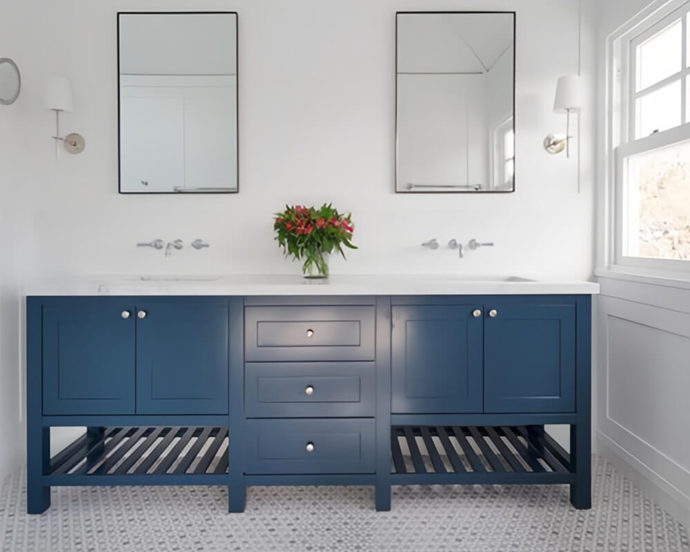Navy blue double-sink bathroom vanity with open shelving, white walls, rectangular mirror, wall sconces, wall-mounted faucets, and red floral arrangement