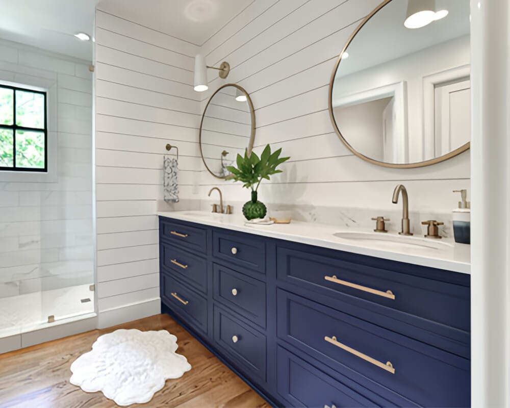 Navy blue double bathroom vanity with marble countertop, brushed gold handles and faucets, wood flooring, gold-framed round mirrors, black-framed shower windows, and greenery