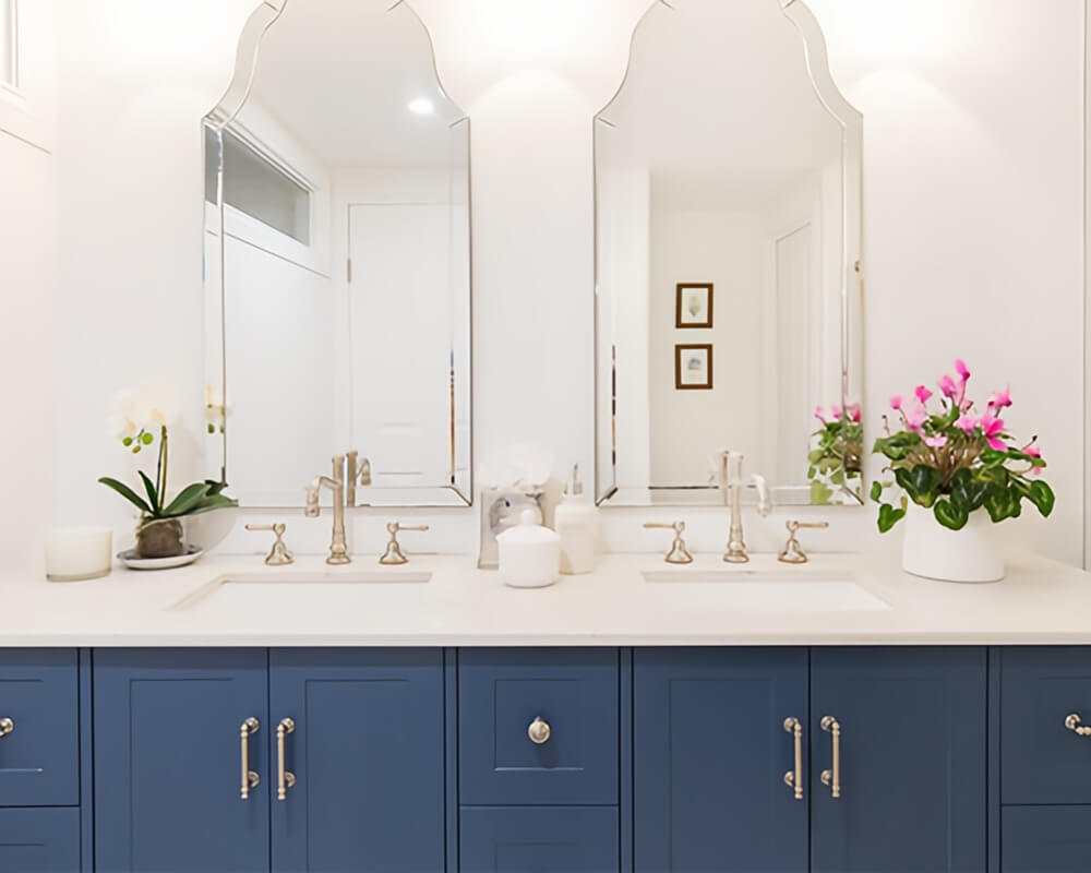 Navy blue double bathroom vanity with white countertop, arched mirrors, brushed gold faucets, wall sconces, brushed nickel handles, greenery, and pink flowers